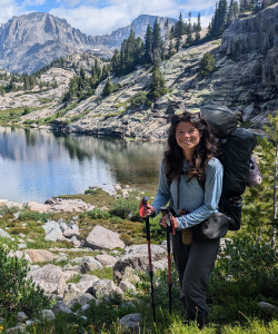 Brynn stands in backpacking gear in front of an alpine lake.