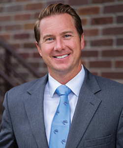 Portrait of Matt standing in front of a brick wall in a dark grey suit. 