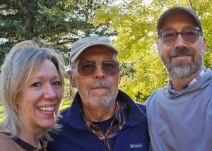 Randy Bruns (center) stands hugging his daughter (left) and son (right) in front of green trees.